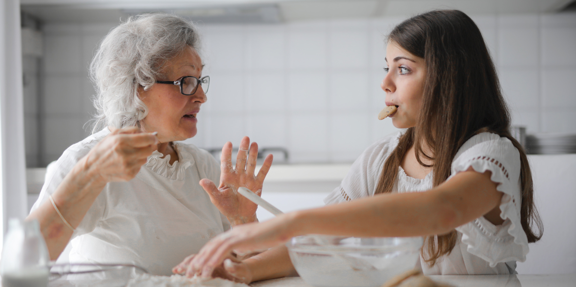 A grandmother sits at a table with her granddaughter baking cookies. The grandmother's hands are covered in flour. They are looking at each other and the girl has a cookie in her mouth.