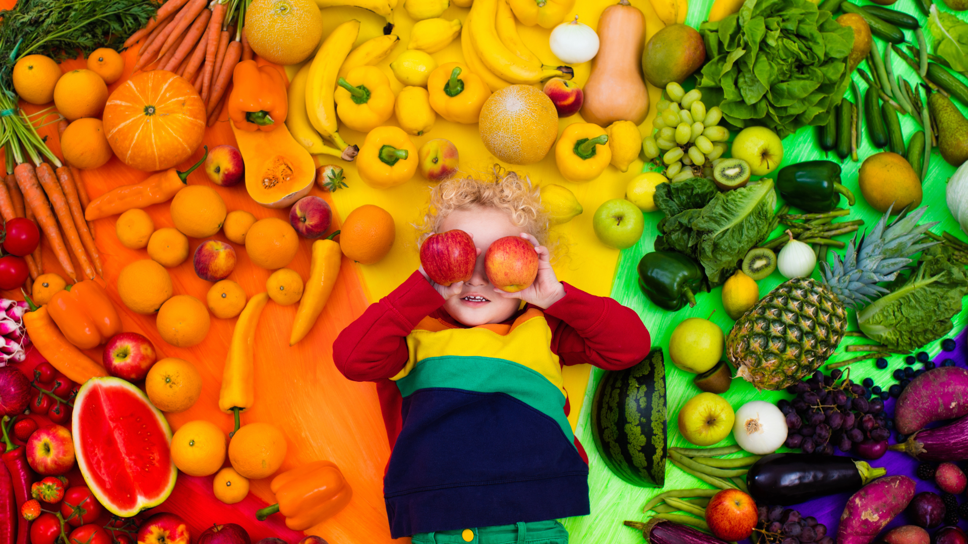 A child sits among an intricate display of produce, Fruits and vegetables are grouped by color, giving the effect of a rainbow. The small child is holding apples up to his face pretending they are his eyes.