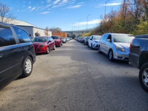 2 lines of cars wait to receive food from The Foodbank's weekly Drive Thru. The line stretches the entire length of the road and wraps around the corner out of view.