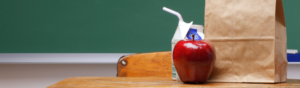 AN apple, milk carton and brown paper lunch bag sit on a wooden school desk, with a blank chalkboard in the background.